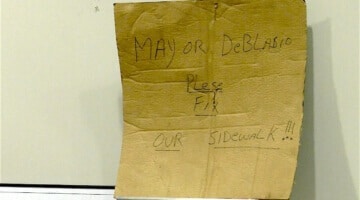 A cardboard sign with hand-written text that reads: "Mayor DeBlasio Please Fix Our Sidewalk!"