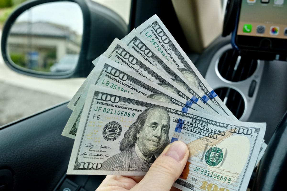 A man's hand holds five $100 bills while seated in a car